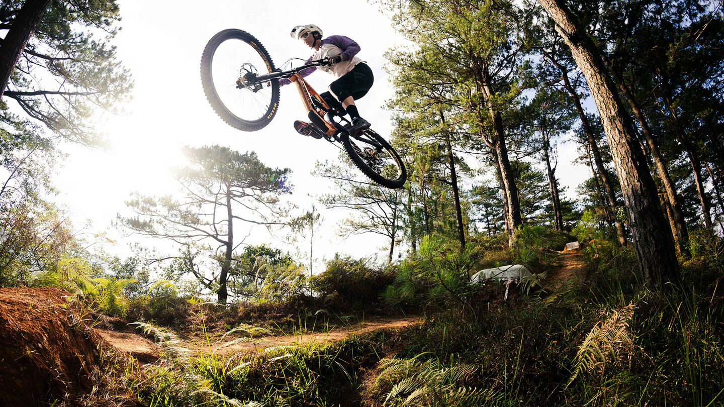 A mountain biker performing a spectacular jump over a hill in a forested area. The rider is wearing protective gear and a helmet, with the sun shining through the trees, bathing the forest in warm light.